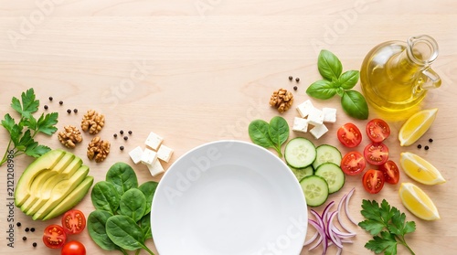 Healthy fresh food ingredients arranged around empty white ceramic plate on light wooden surface including avocado spinach tomatoes cucumber feta cheese and oil.