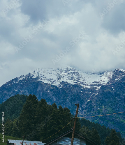 Fotografía : A dramatic snow-covered mountain peak partly wrapped in cloud, viewed above a dense pine forest and a rustic tin-roofed cabin
