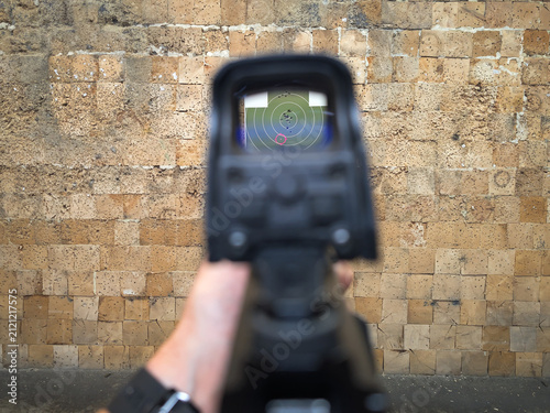 Obraz na płótnie First-person point of view aiming rifle through holographic sight reticle at green target on wooden wall in indoor shooting range