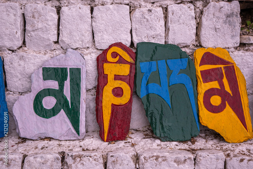 Fotografie Brightly colored stones engraved with the mantra “Om mani padme hum,” arranged in front of a wall of white stones