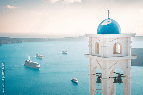 White church bell tower with blue dome overlooking deep blue caldera and cruise ships in santorini, greece