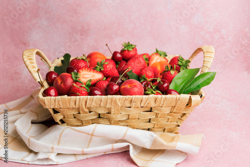 Fresh strawberries and cherries, aprico  in a woven basket on a light pink background with a dish towel