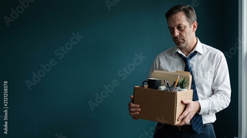 A dejected middle-aged man in a suit holding a cardboard box filled with office supplies after being laid off.