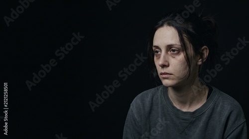 A young woman with dark circles under her eyes looks off to the side with a somber expression against a black background.