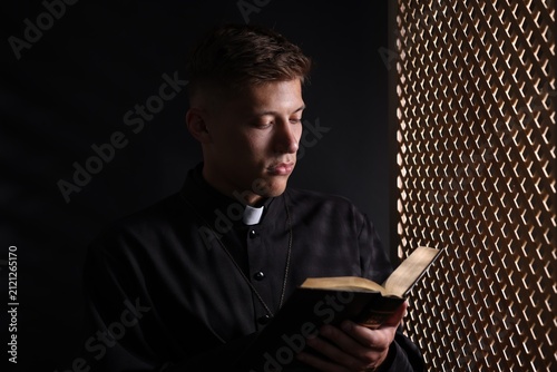 Priest with Holy Bible praying near partition in confessional booth