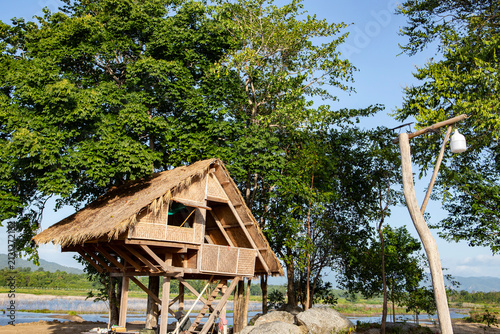 Fotografie A traditional rustic bamboo hut with a thatched roof elevated on stilts, surrounded by lush green trees near a scenic lake under a clear blue sky