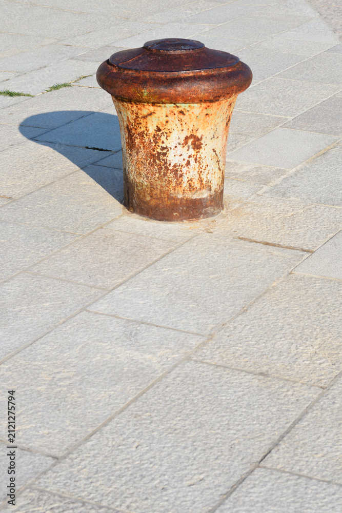 Naklejka premium A single old rusty iron mooring bollard stands firmly on a paved stone pier under bright natural sunlight