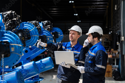 Concept of Industry 4.0 with professional engineers collaborating on automation technology, using laptop and tablet to program blue robotic arm in a modern smart manufacturing factory.