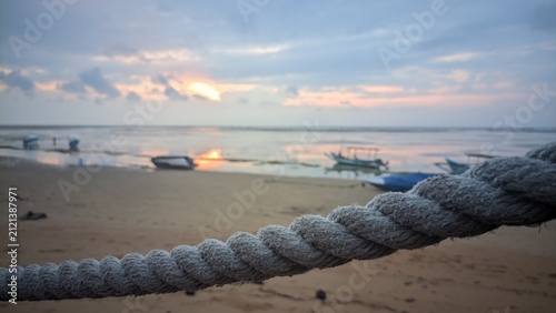 Close up of a thick weathered nautical rope with a blurred tropical beach sunset and traditional fishing boats in the background.