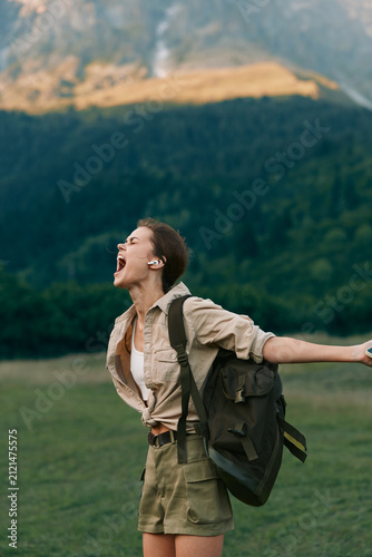Lively woman outdoors, laughing with mouth open, backpack on shoulder, wide grassy field, mountains in the background, sunlit moment, carefree lifestyle