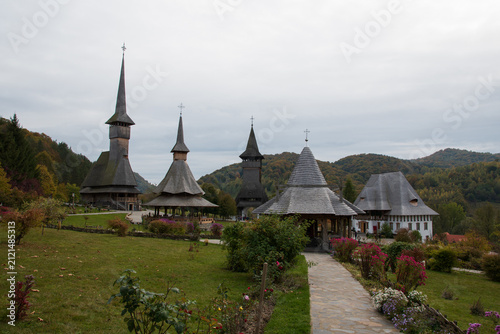 Historic wooden church buildings with steep shingle roofs, flower gardens, and forested hills beneath an overcast autumn sky.