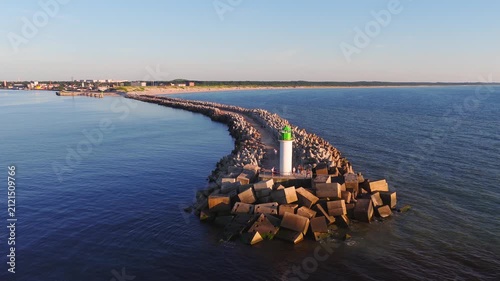 Aerial view of Ventspils, Latvia, shows curved tetrapod breakwater to a white lighthouse with green lantern, sandy beach, woods, port tanks, and people on the pier.