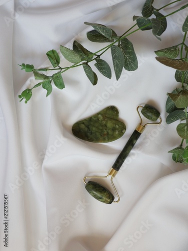 Obraz na plátne Green jade facial massage roller and heart-shaped gua sha stone arranged on soft draped white fabric beneath a eucalyptus branch