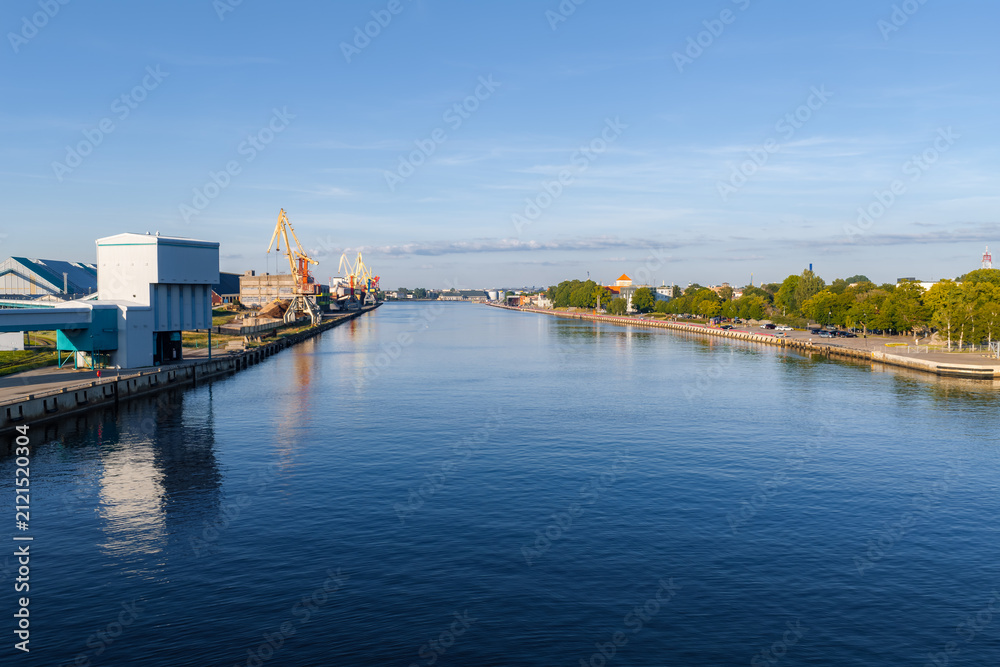 Obraz premium Wide channel toward the Port of Ventspils in Latvia, cargo cranes and terminals on the left, a promenade on the right, calm water in morning light, modern buildings and silos.