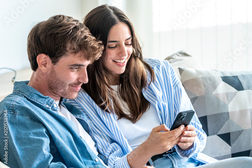 Young couple sitting comfortably on a sofa at home  smiling while sharing and looking at content on a mobile phone  representing connection  leisure  and modern lifestyle