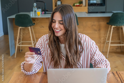 Happy young woman making an online payment using her credit card and laptop while sitting at a kitchen counter, enjoying the convenience of e-commerce and digital finance