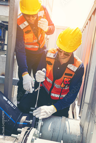 Two industrial technicians in safety gear repairing a pipeline system at a factory. Construction workers in hard hats and reflective vests fixing large water pipesSupervisor with walkie-talkie monitor