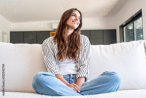Принти на полотні Woman feeling happy and comfortable, sitting cross-legged on a white couch in a