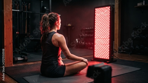 Female Athlete Experiencing Full Body Red Light Therapy Panel Session In Dark Home Gym For Muscle Recovery