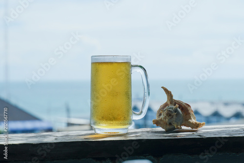 Chilled beer mug on the blue wooden table. Blurred sea is at the background. Cooled glass of beer on the wooden table.