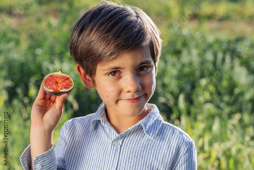 Smiling little boy holding freshly cut ripe fig slice while looking at camera. Healthy childhood, natural nutrition, seasonal fruit, organic harvest and joyful family lifestyle.