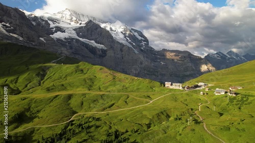 Wide panoramic view of Kleine Scheidegg in Swiss Alps with isolated chalets against massive snowy mountain peaks in soft morning light. Sunrise over green alpine meadows. Bernese Oberland Switzerland