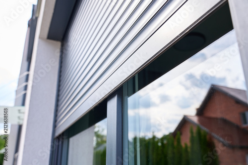 Partially lowered metal roller shutter covers reflective exterior window showing nearby houses and cloudy sky