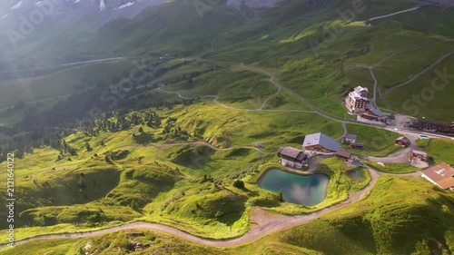 Aerial drone view of Kleine Scheidegg in Swiss Alps with green meadows and snowy Eiger Monch peaks in early morning light. Small station with emerald lake and winding road in 4K video
