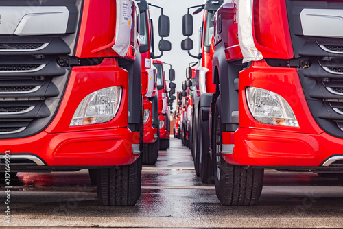 Close up of new red commercial semi truck cabins with headlights and tires parked in rows at logistics terminal