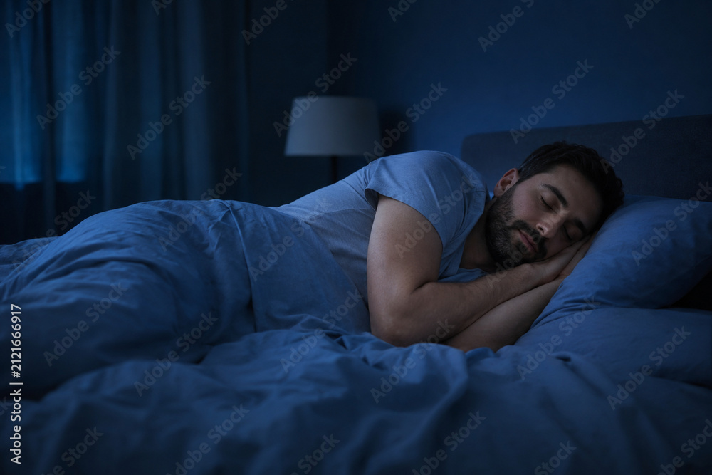 Fototapeta premium Man Sleeping Peacefully: A man rests soundly in a dark bedroom, nestled under blue comforters, with a soft lamp illuminating the quiet space. Evokes a sense of calm and rest.