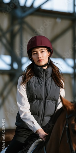 Young female equestrian portrait riding horse indoors wearing burgundy helmet and gray vest for lifestyle banner background