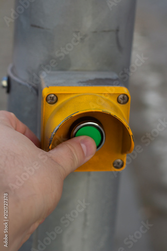 Vertical street photo. Finger presses green button in yellow case. Device for switching traffic light signal on pedestrian crossing on metal pole. Concept of safety, regulation of people movement