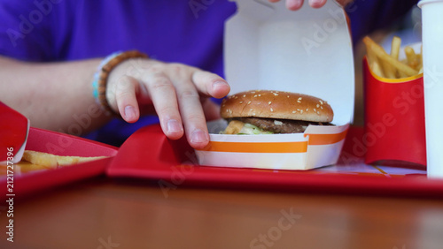Defocused mobile photo with selective focus close up. Fat man sits at table of fast food cafe with French fries on tray and takes burger from white paper box. Male hand with blue t-shirt, bracelet