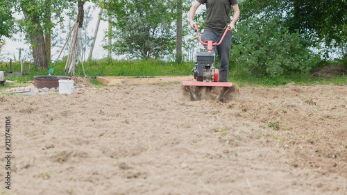 A young Caucasian man operates a tiller in a garden. The soil is freshly turned, surrounded by greenery and trees in the background.