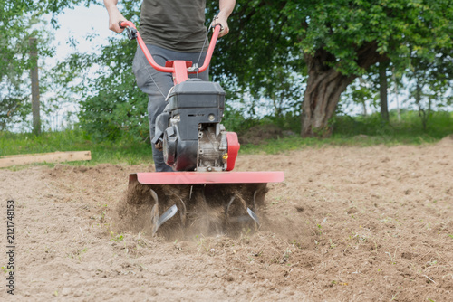 A man operates a red tiller in a field. The ground is being turned up, creating dust. Green trees are visible in the background.