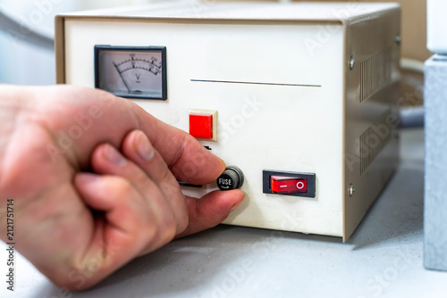 Close-up of a human hand pressing a red rocker power switch on a electrical control unit in a workshop or laboratory setting.