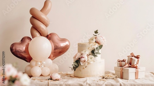 Cake decorated with soft floral arrangements sits beside metallic balloons and wrapped gift boxes on a light table.