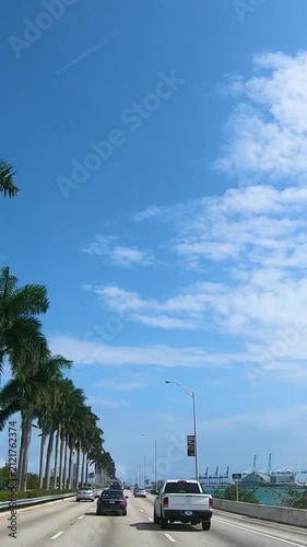 Vertical POV drive along a Miami freeway in Florida with moving traffic, palm trees and a modern urban atmosphere on a sunny day.