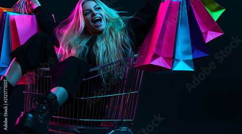 young blonde woman riding inside a metal shopping cart, holding up colorful paper shopping bags