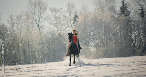Woman riding black horse through snow in winter forest