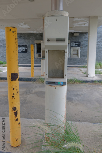 An abandoned bank drive-through with a closed ATM and teller window set in a weathered brick facade, featuring peeling ceiling, stained concrete and overgrown weeds that convey neglect and urban decay