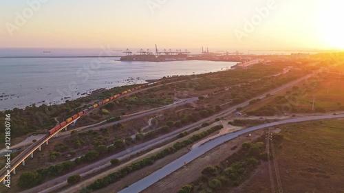 Freight train traveling on a bridge along the coastline near the Port of Sines at sunset in Sines, Setubal, Portugal. Aerial view. Moving forward 