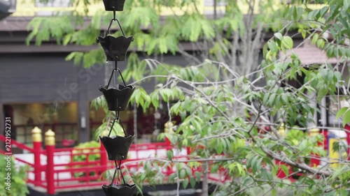 Traditional Japanese metal rain chain kusari doi with decorative cup design in garden setting. Rain water flows through the cups against blurred background of green trees and red bridge.