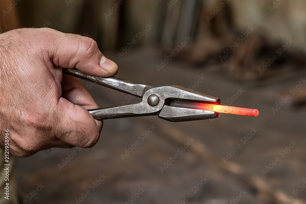 Obraz premium Blacksmith's hand holding glowing hot metal rod with sturdy tongs in workshop