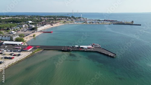 Aerial footage over the historic Mackinac City shoreline. Piers and docks jut from the sandy shoreline for the tour boats to moor. City and bridge alive with adventure and experience