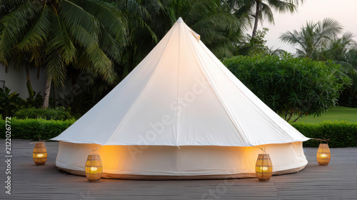 A white canvas bell tent illuminated by warm lights in a tropical setting. Tall palm trees and lush greenery surround the tent, creating a serene atmosphere