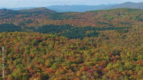 AERIAL: Winding road curves through densely forested hills covered in green, yellow and orange autumn foliage near Otočac, Croatia. Expansive landscape in colors of fall with distant blue mountains.