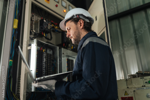 Fotografía Electrical engineer using laptop near open electrical cabinet, conducting control system diagnostics, facility maintenance management, workplace safety compliance