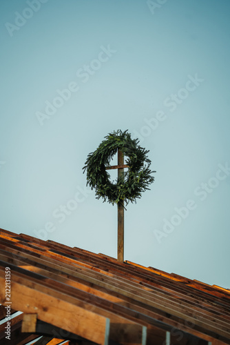 Close-up of a traditional evergreen topping-out wreath mounted above a timber house under construction against a clear blue sky.