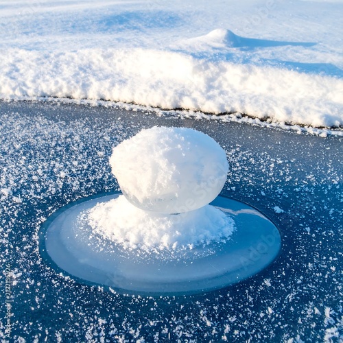 Round Snowball Resting on Thin Ice Disk on Frozen Lake Surface